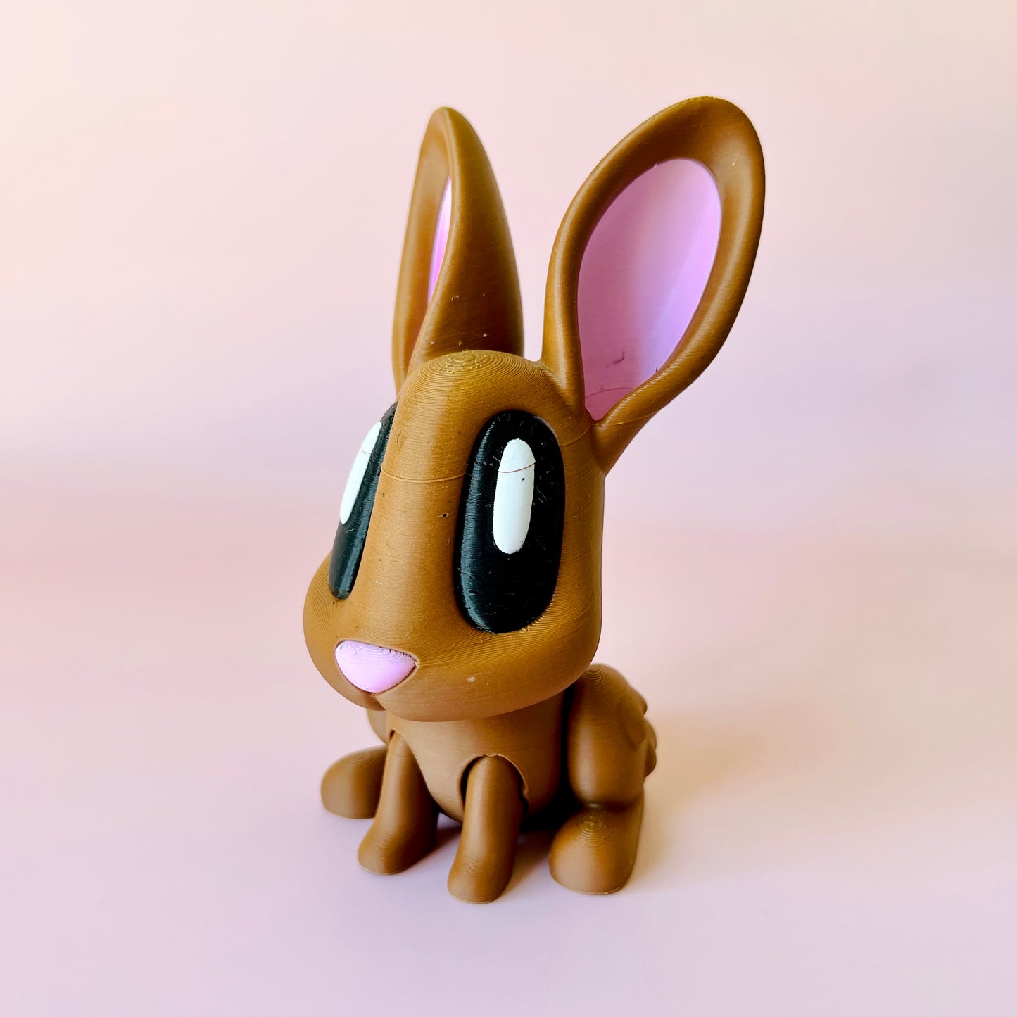 A cute brown rabbit figure with large black eyes and pink ears, set against a soft pink background.