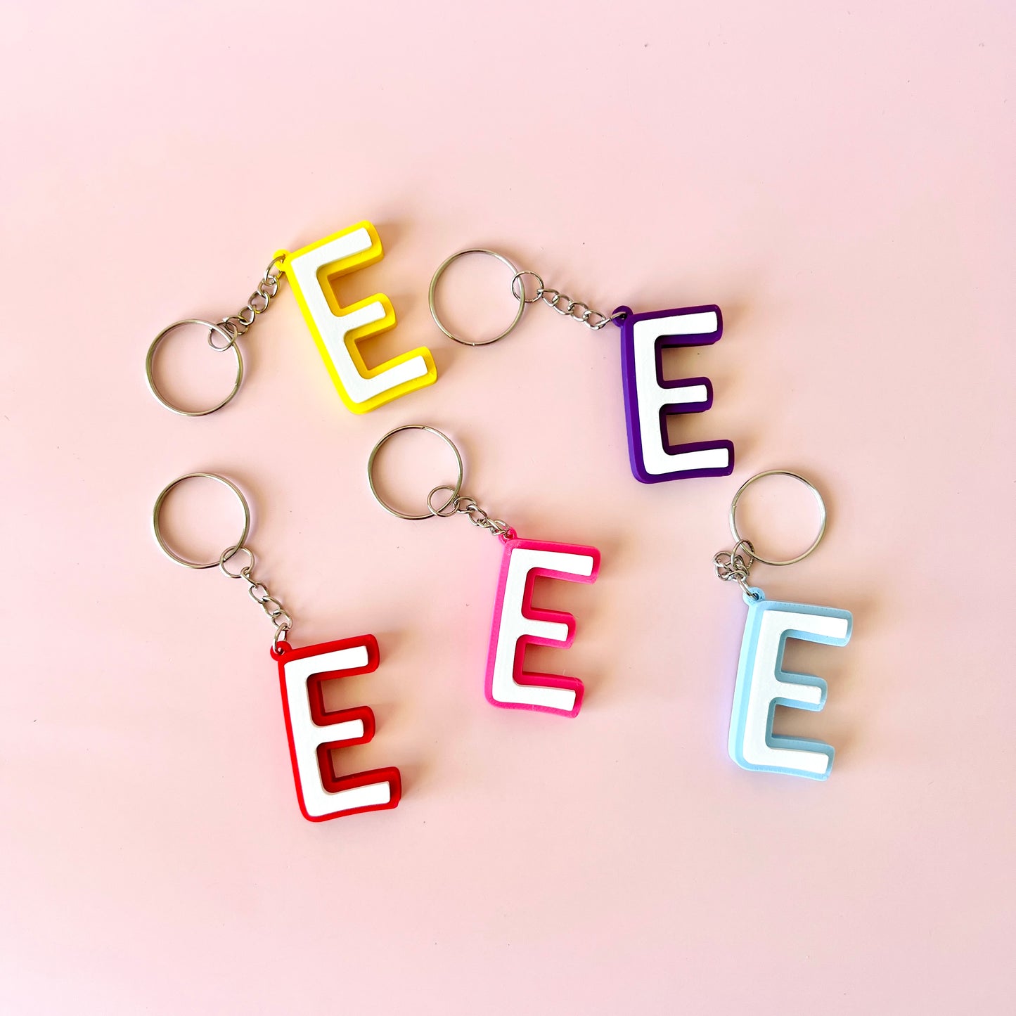 Colorful letter "E" keychains in yellow, red, pink, purple, and blue against a soft pink background.