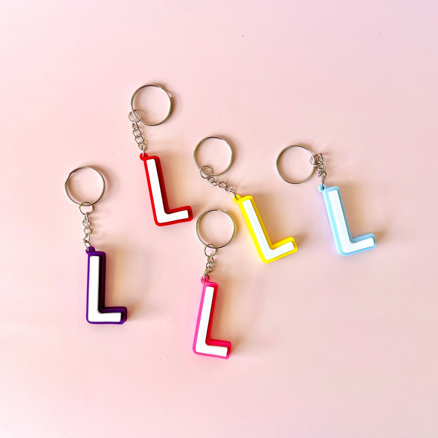 Five colorful keychains featuring the letter "L" in various shades on a soft pink background.