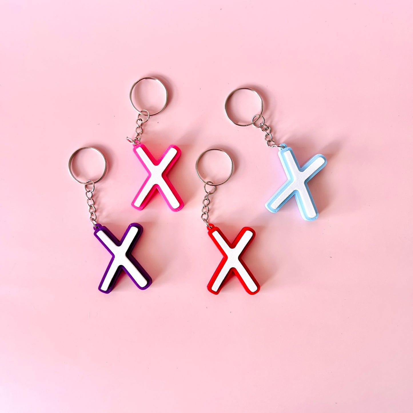 Colorful keychains in pink, purple, red, and blue shaped like the letter "X" on a soft pink background.