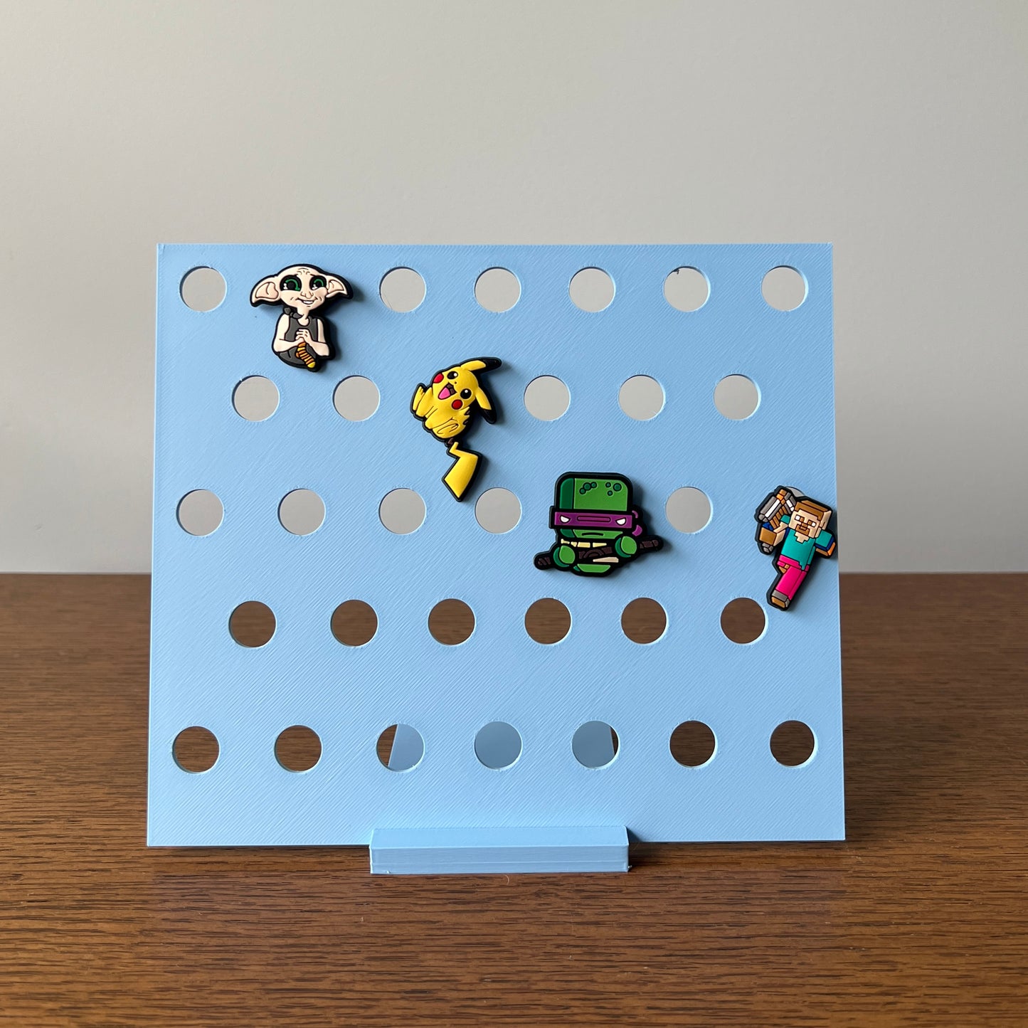 A light blue display stand showcasing colorful character charms with circular cutouts for easy arrangement and visibility.
