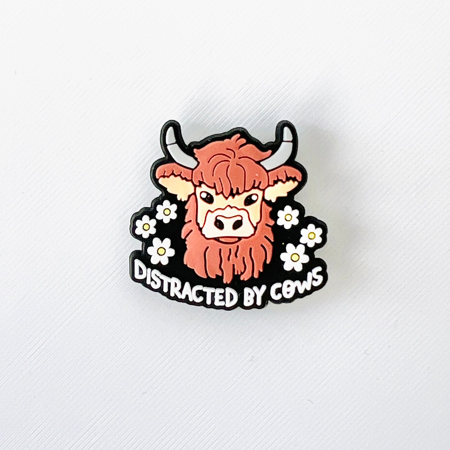 Brown Highland cow surrounded by white flowers with the phrase "Distracted by Cows" for a fun, quirky shoe charm.
