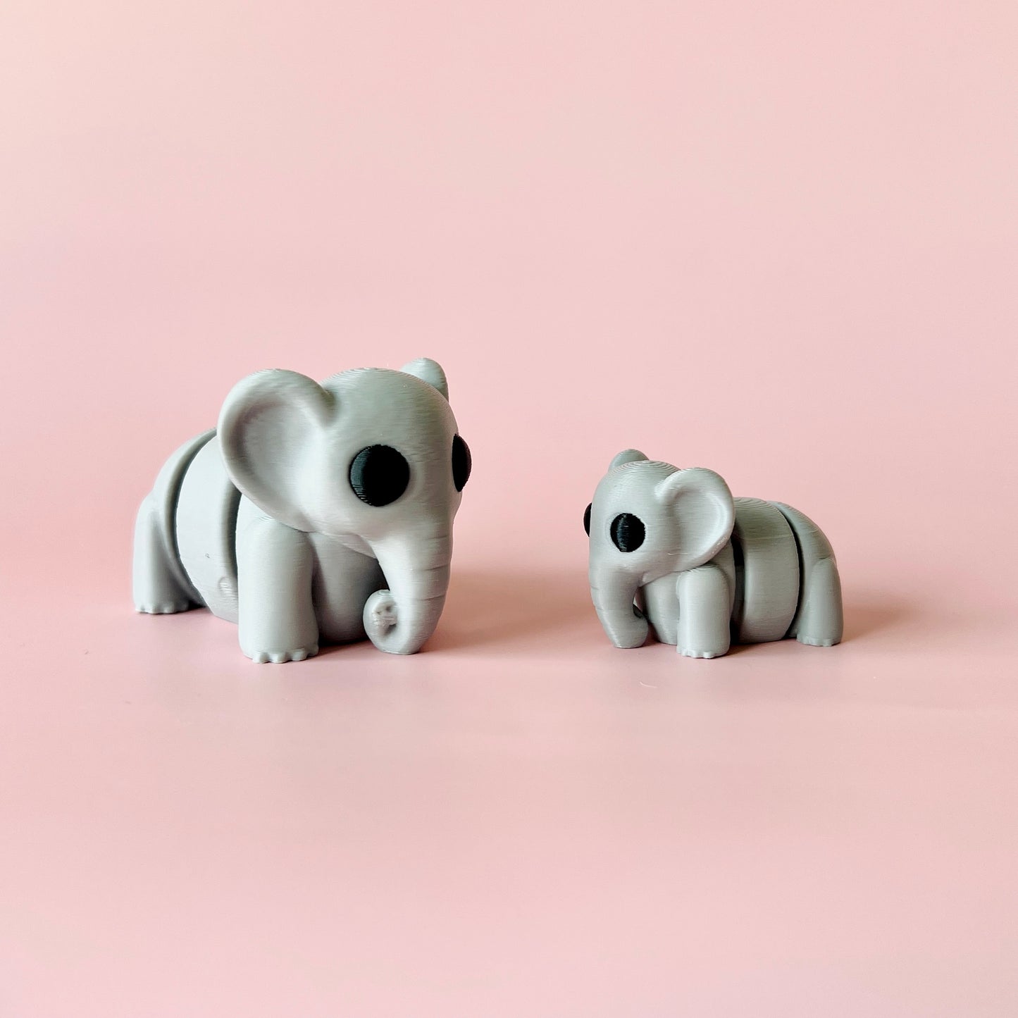 Two stylized gray elephants with black eyes, positioned on a soft pink background, showcasing their charming features.