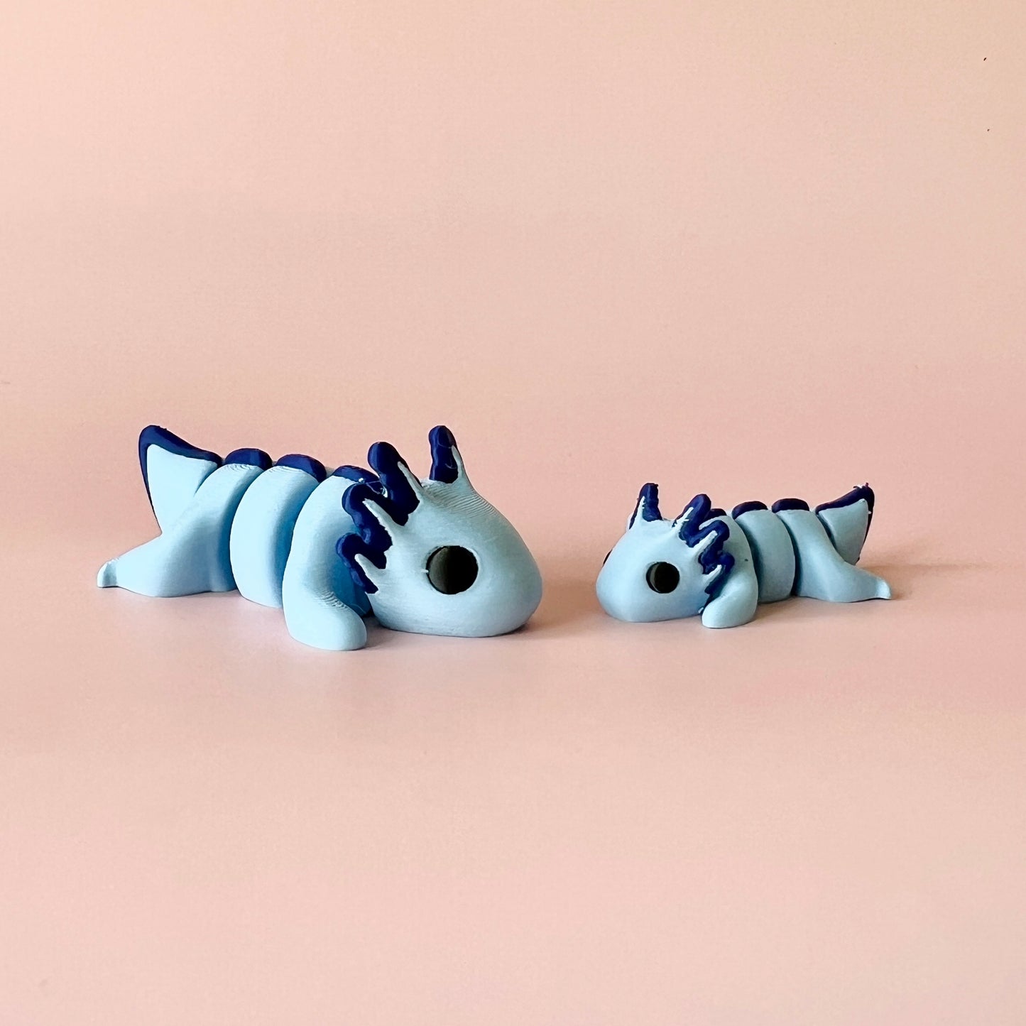 Two charming blue creatures with dark blue accents, resting on a soft pink background, showcasing playful, whimsical shapes.