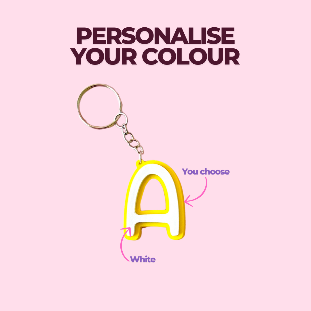 A vibrant yellow keychain in the shape of the letter "A" with a customizable color option, against a pink background.