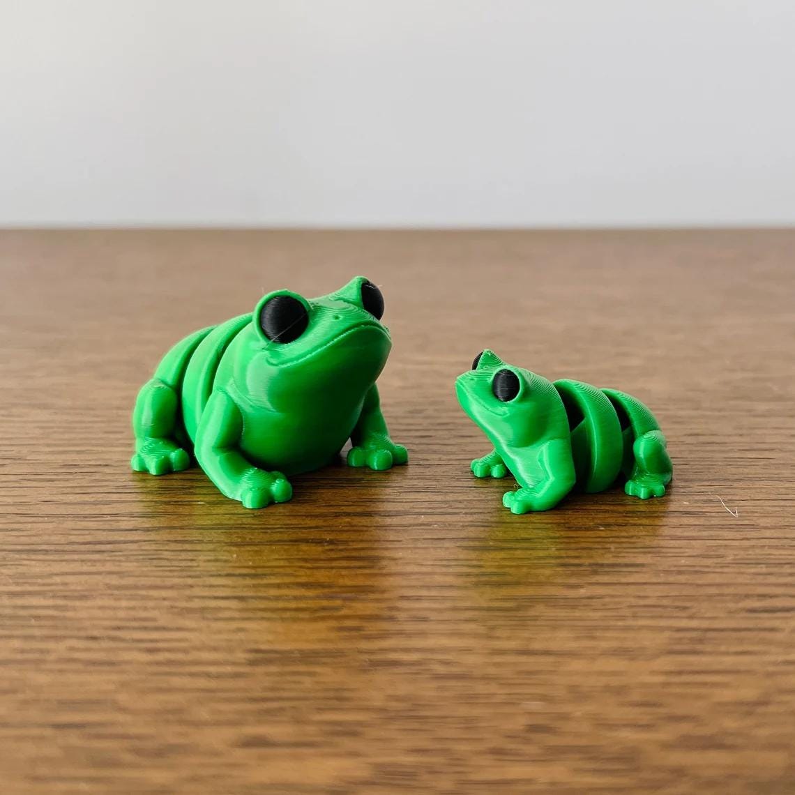 Two vibrant green articulated frogs, one larger with a smooth body and the other smaller, both featuring black eyes.