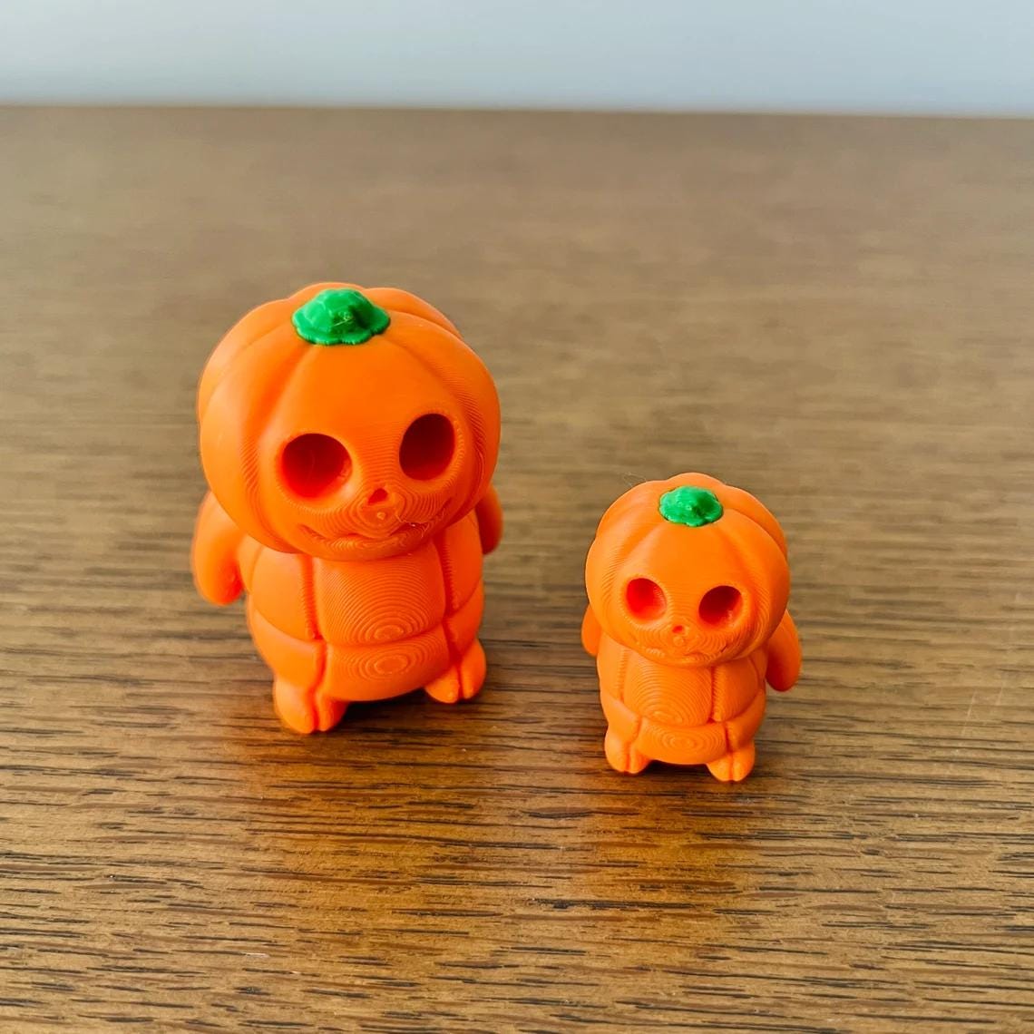 Two cute articulated pumpkins in bright orange, one larger with a green stem and a smaller version, on a wooden surface.