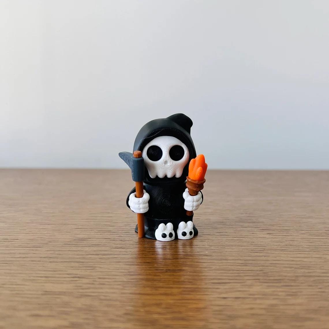 Adorable mini Grim Reaper holds a scythe and torch, featuring black cloak and cute bunny slippers, perfect for collectibles.