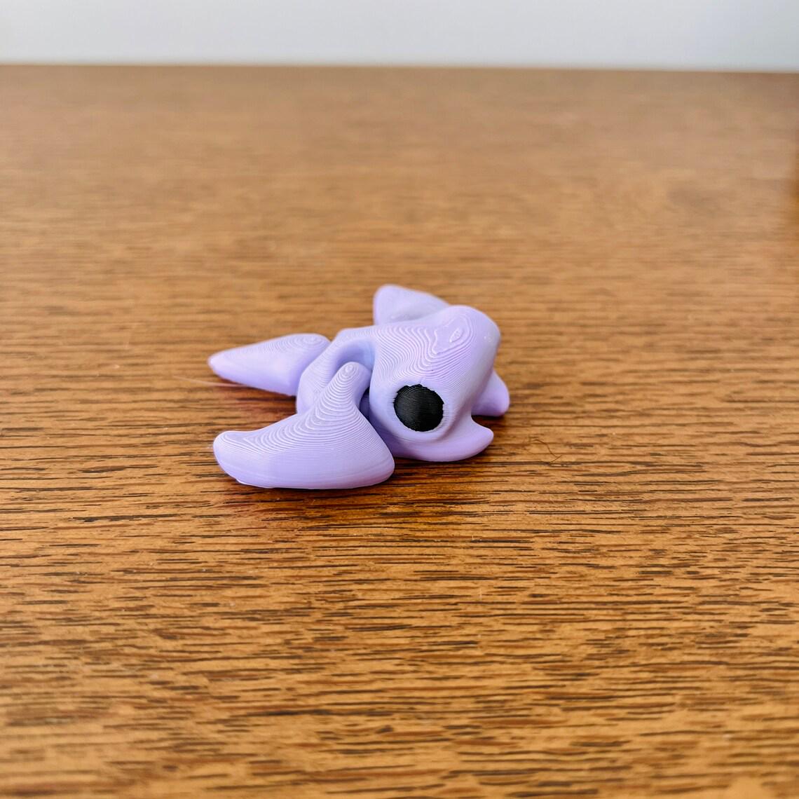 Articulated mini stingray in soft lavender with a black eye, resting on a wooden surface for sensory play and stress relief.
