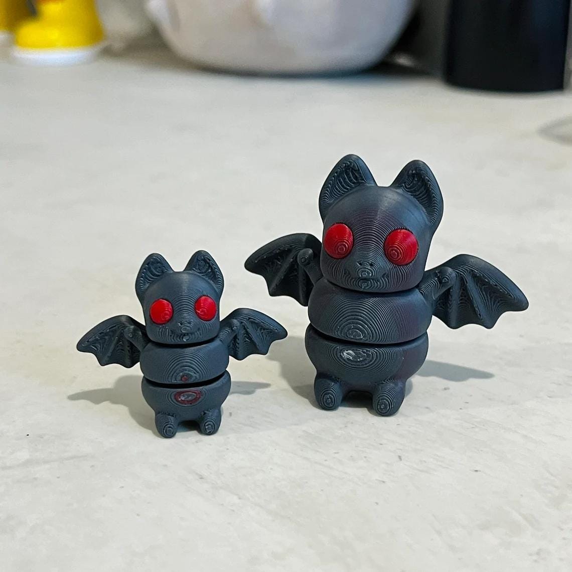 Two 3D printed bats, one small and one large, with red eyes and articulated wings, resting on a light surface.
