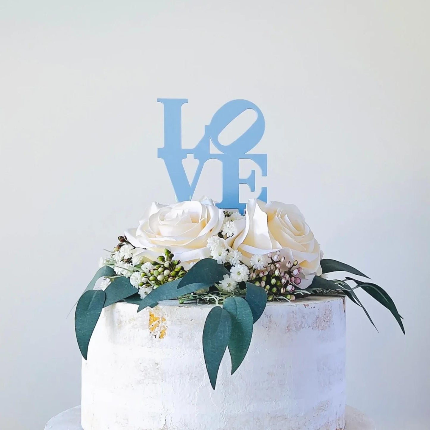 A light blue "LOVE" topper sits atop a decorated cake with white roses and green foliage, adding a romantic touch.