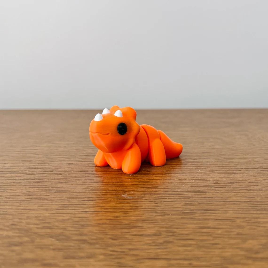 Cute orange Mini Triceratops with black eyes and white horns, resting on a wooden surface. Perfect for sensory play!