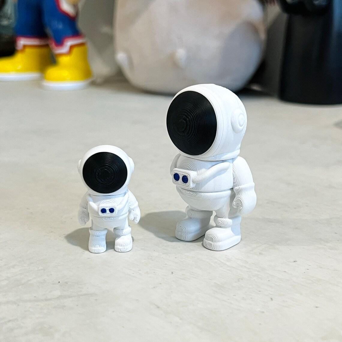Two articulated astronauts in white with black helmets, one small and one larger, on a textured surface.