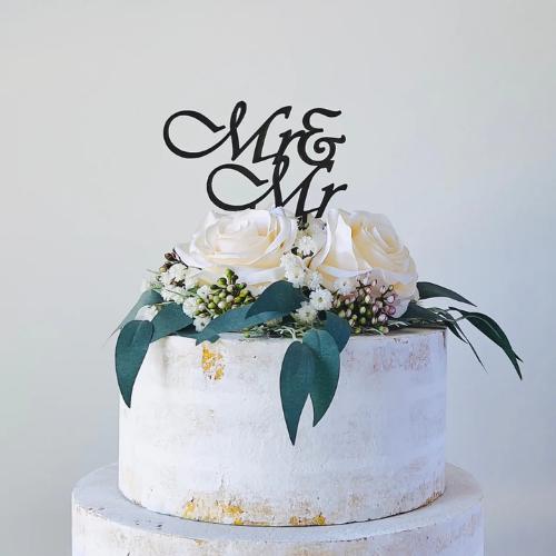 Elegant black "Mr & Mr" topper above white roses and greenery on a rustic wedding cake, adding charm to the celebration.
