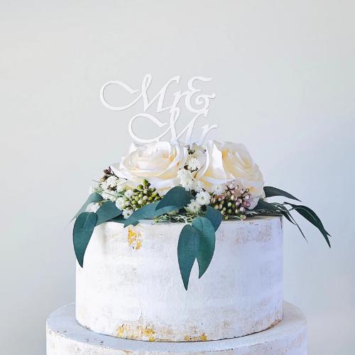 Stylish white "Mr & Mr" topper atop a decorated cake with soft roses and greenery, perfect for a wedding celebration.