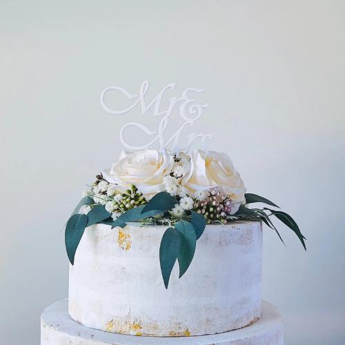 Elegant "Mr & Mrs" topper on a cake adorned with white roses and greenery, perfect for a wedding celebration.