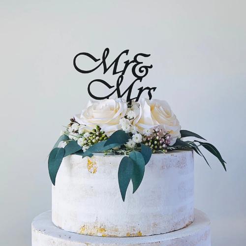 Elegant black "Mr & Mrs" topper atop a white cake adorned with white roses and greenery, perfect for weddings.