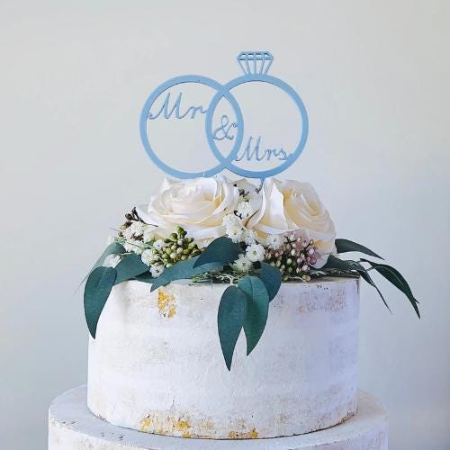Interlocking rings with "Mr & Mrs" and a diamond accent atop a floral-adorned wedding cake, exuding romance and elegance.