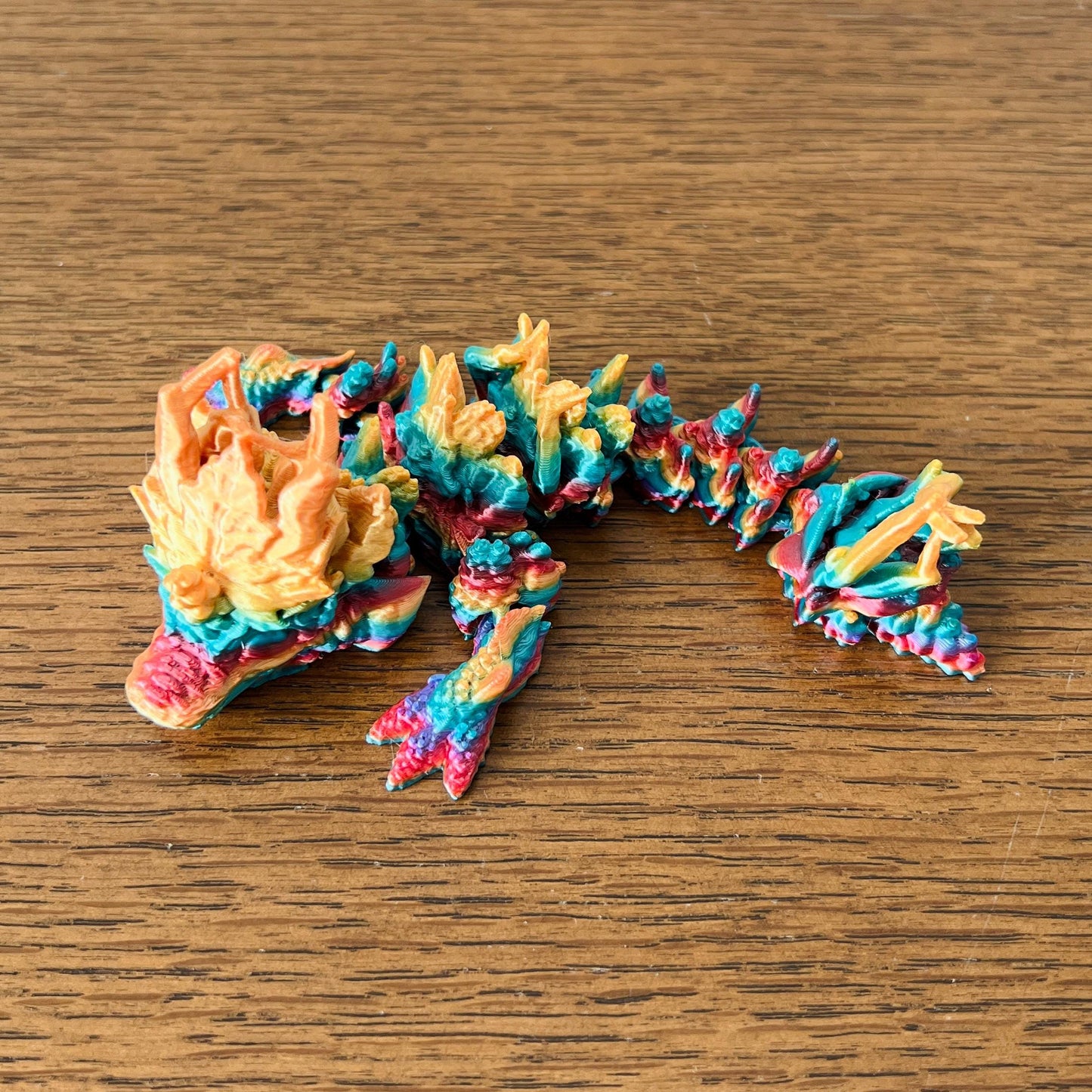 Colorful wingless dragon with intricate autumn leaf textures in shades of orange, red, and teal, resting on a wooden surface.