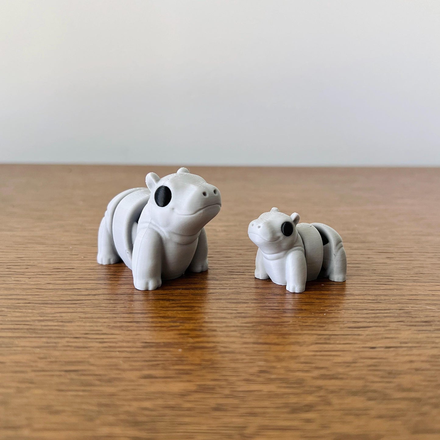 Two adorable mini hippos in gray, featuring articulated bodies and playful expressions, resting on a wooden surface.