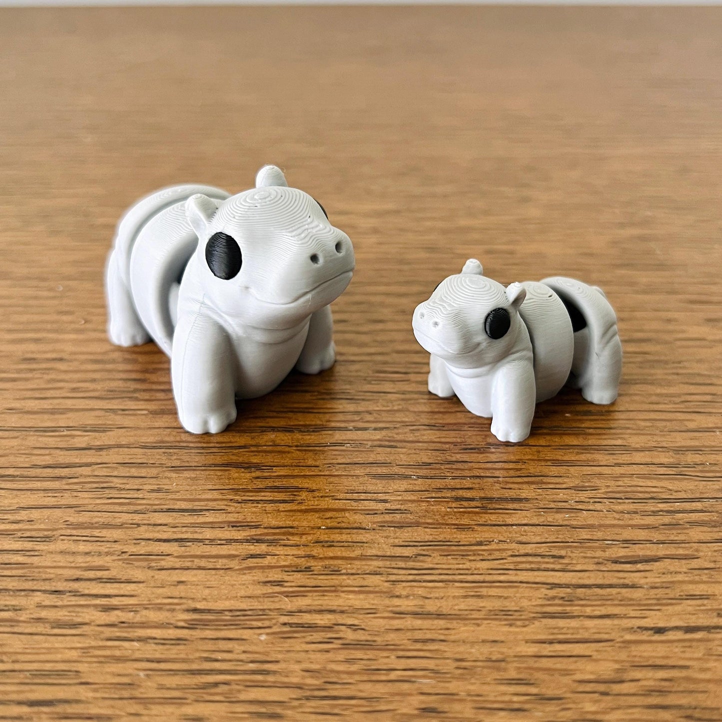 Two cute, articulated mini hippos in gray, showcasing playful expressions and black accents on a wooden surface.