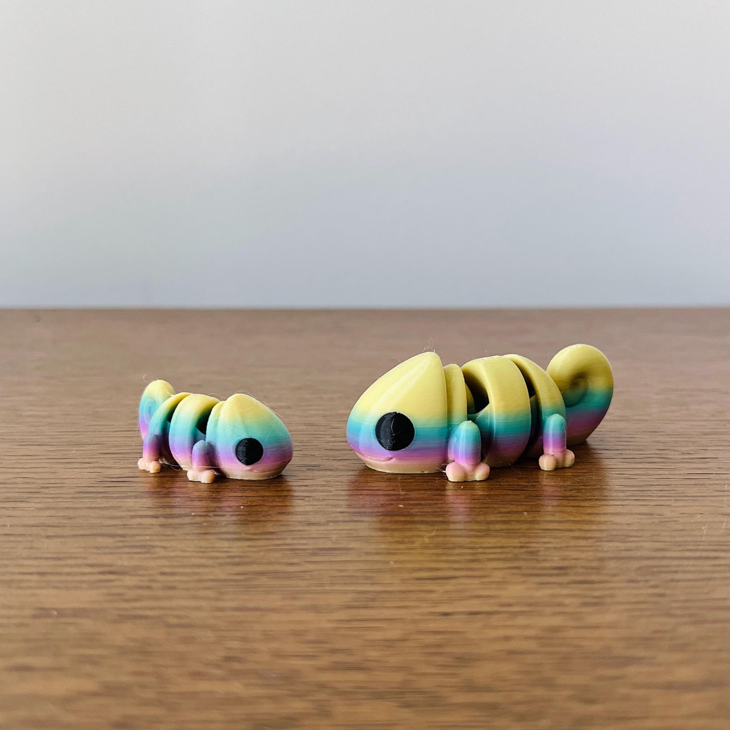 Two articulated chameleons in vibrant rainbow colors, one larger and one smaller, on a wooden surface.
