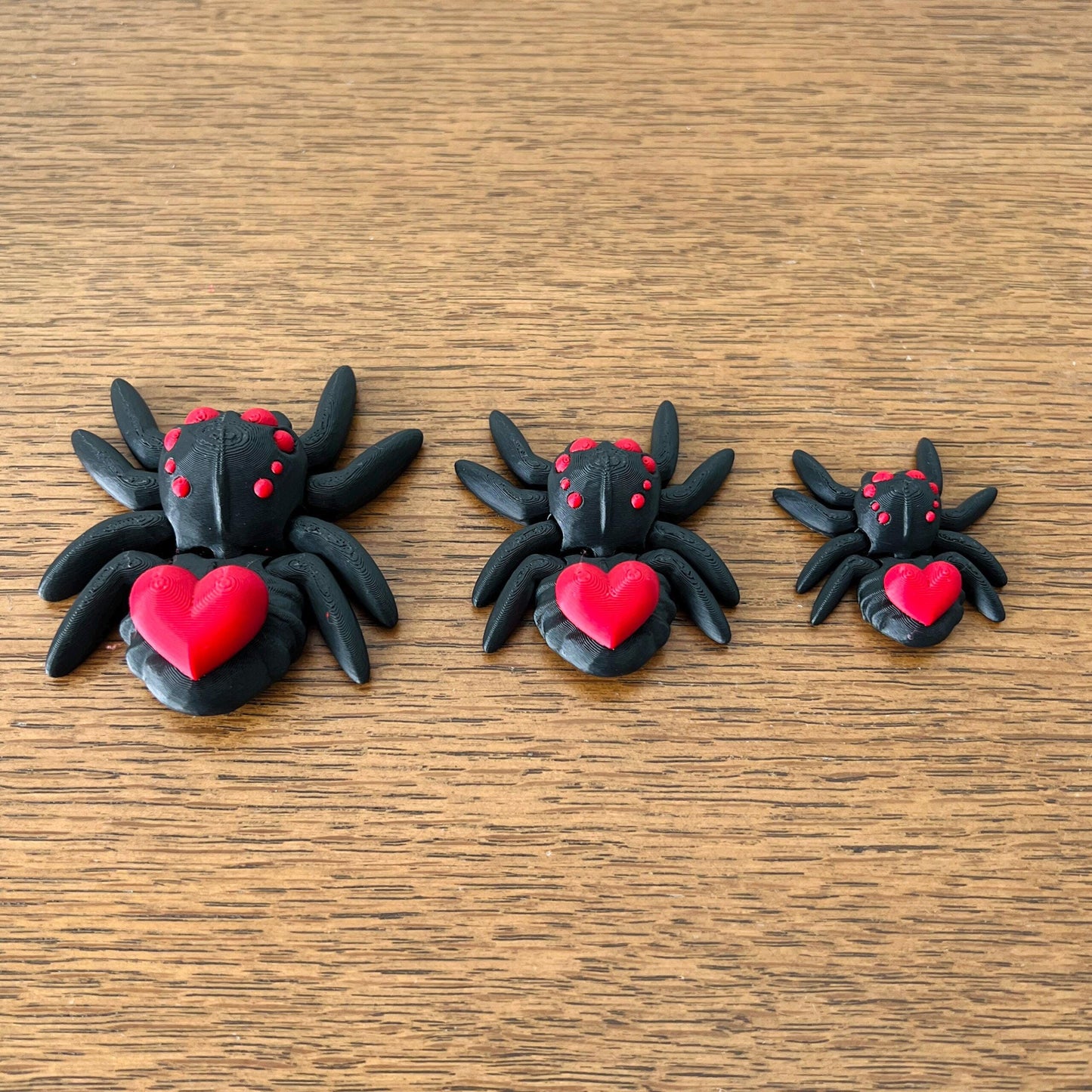 Three adorable black spiders with red hearts on their backs, varying in size, resting on a wooden surface.