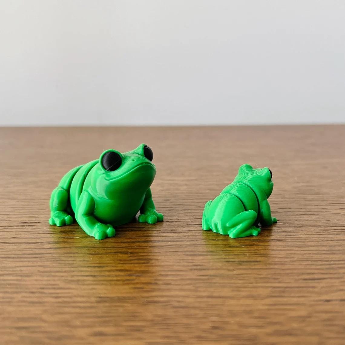 Two green articulated frogs with large eyes, one larger and one smaller, resting on a wooden surface.