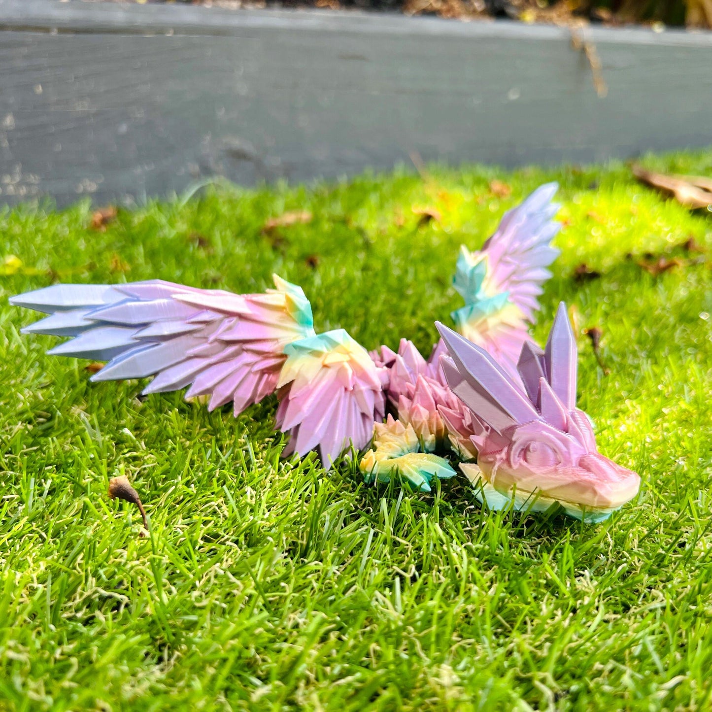 3D-printed dragon with crystal-like pastel wings, resting on green grass, showcasing a whimsical fantasy charm.