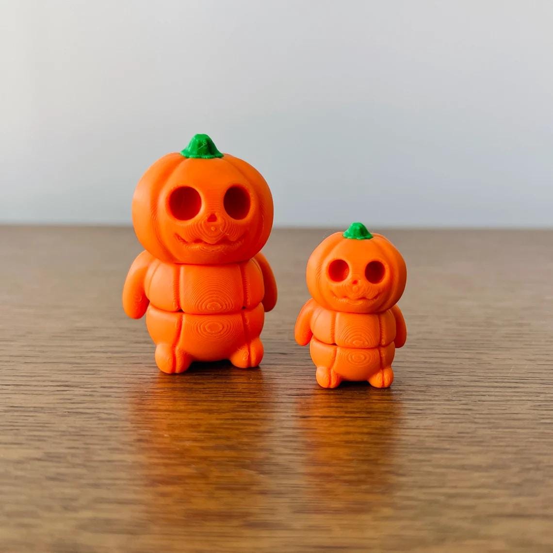 Two cute, articulated orange pumpkins with green tops, featuring playful expressions, on a wooden surface.
