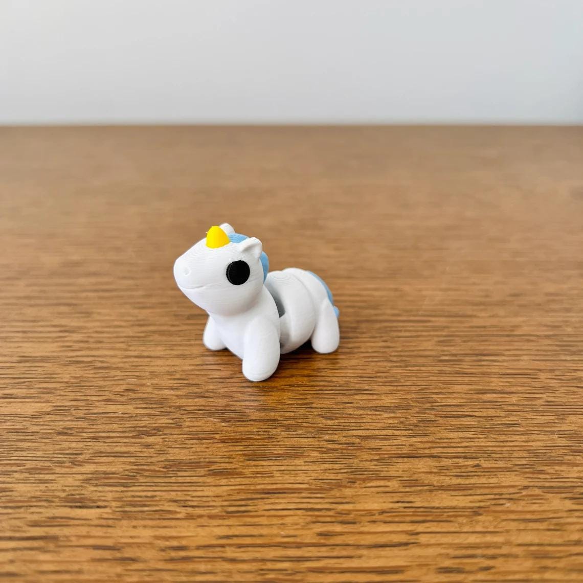 A small, white unicorn with blue mane and yellow horn, resting on a wooden surface, radiating charm and whimsy.
