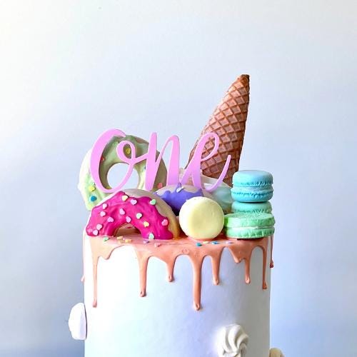 Playful cake topper spells out "ONE" in pink, adorned with colorful macarons, donuts, and an ice cream cone. Perfect for firs