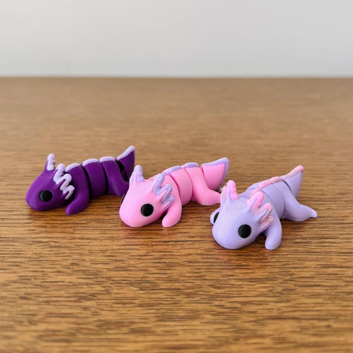 Pastel purple axolotl with articulated limbs, alongside pink and purple counterparts, on a wooden surface.