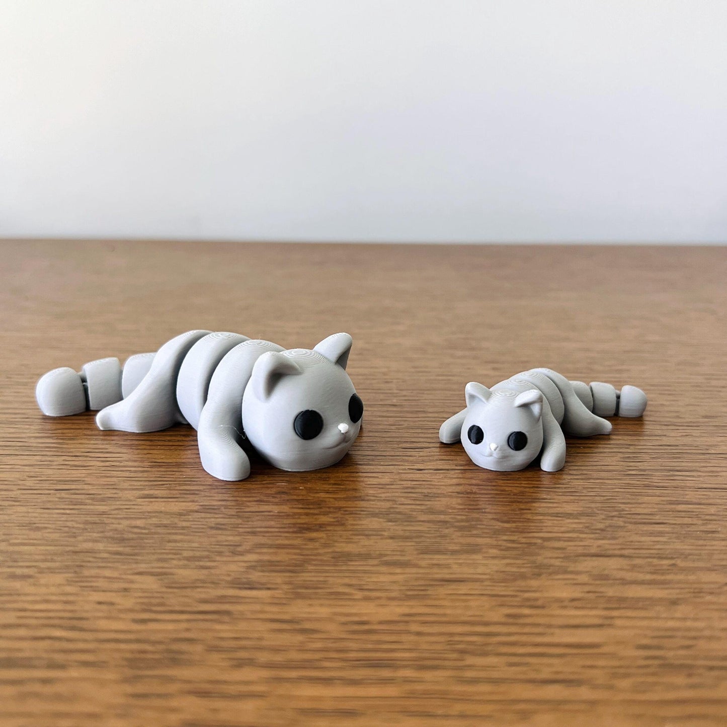 Two adorable articulated grey cats, one small and one slightly larger, resting on a wooden surface.