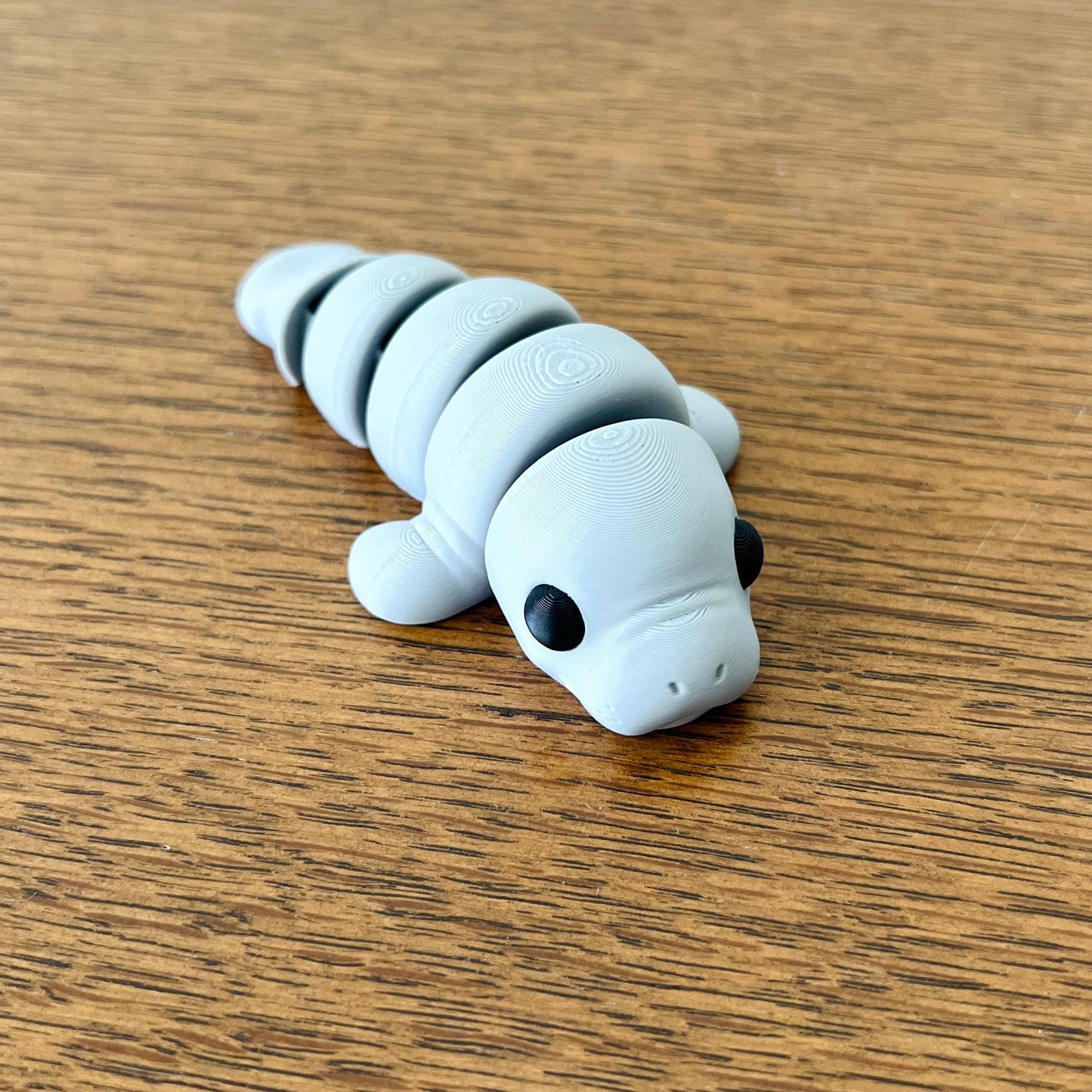 Adorable gray mini manatee with articulated segments and large black eyes, resting on a wooden surface.