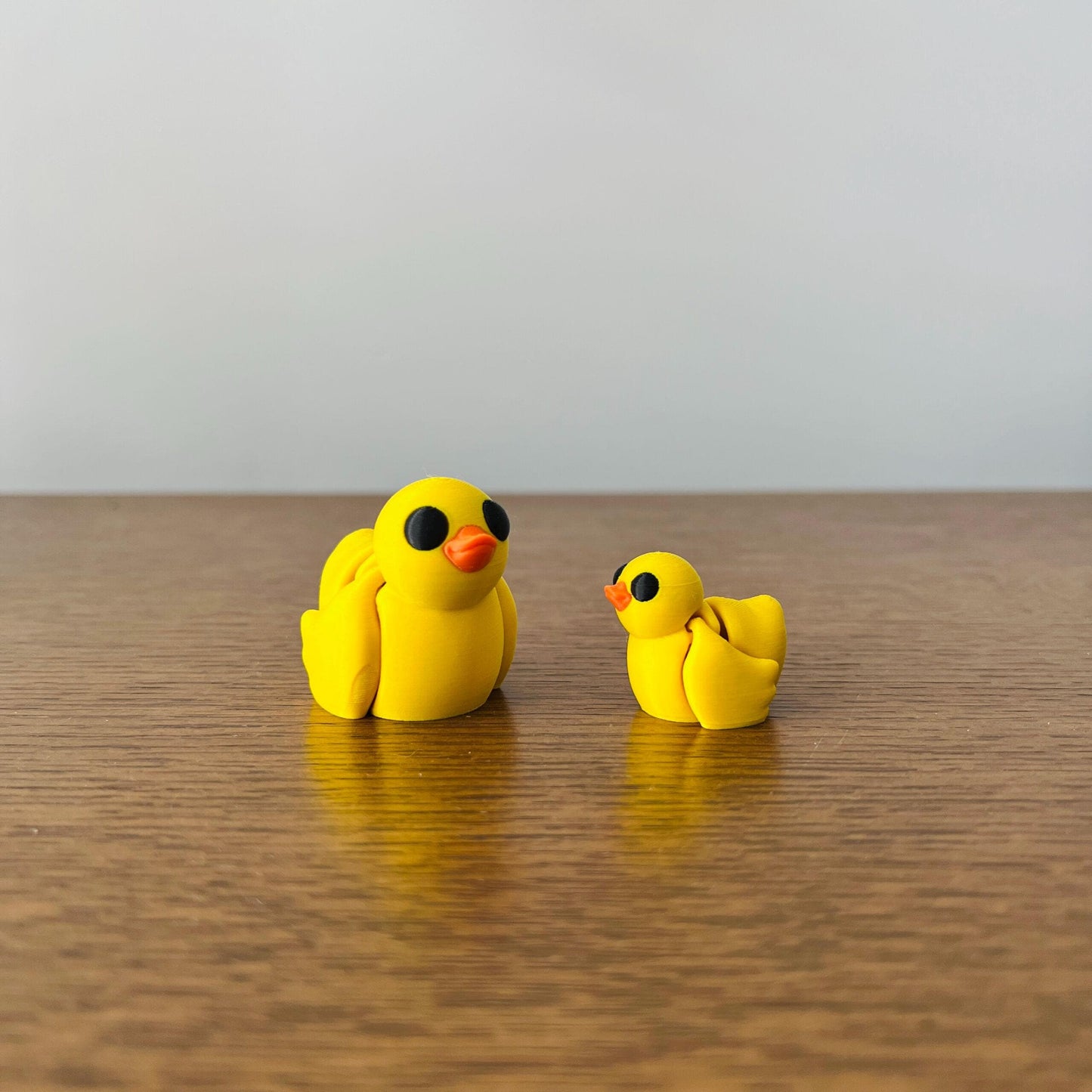 Two adorable, bright yellow ducks with big black eyes, one larger and one smaller, sitting on a wooden surface.