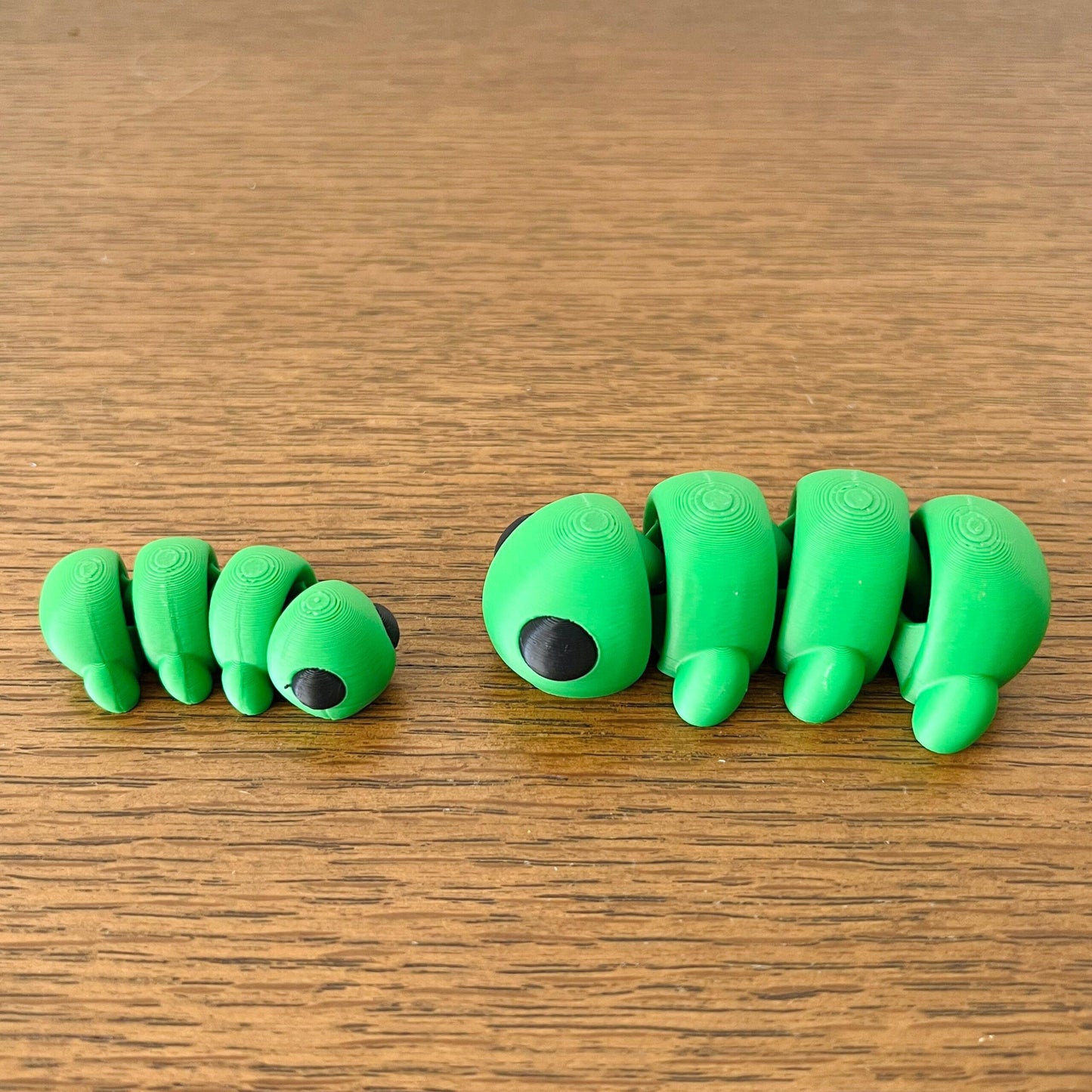 Two playful green caterpillars with black eyes, one smaller than the other, resting on a wooden surface.