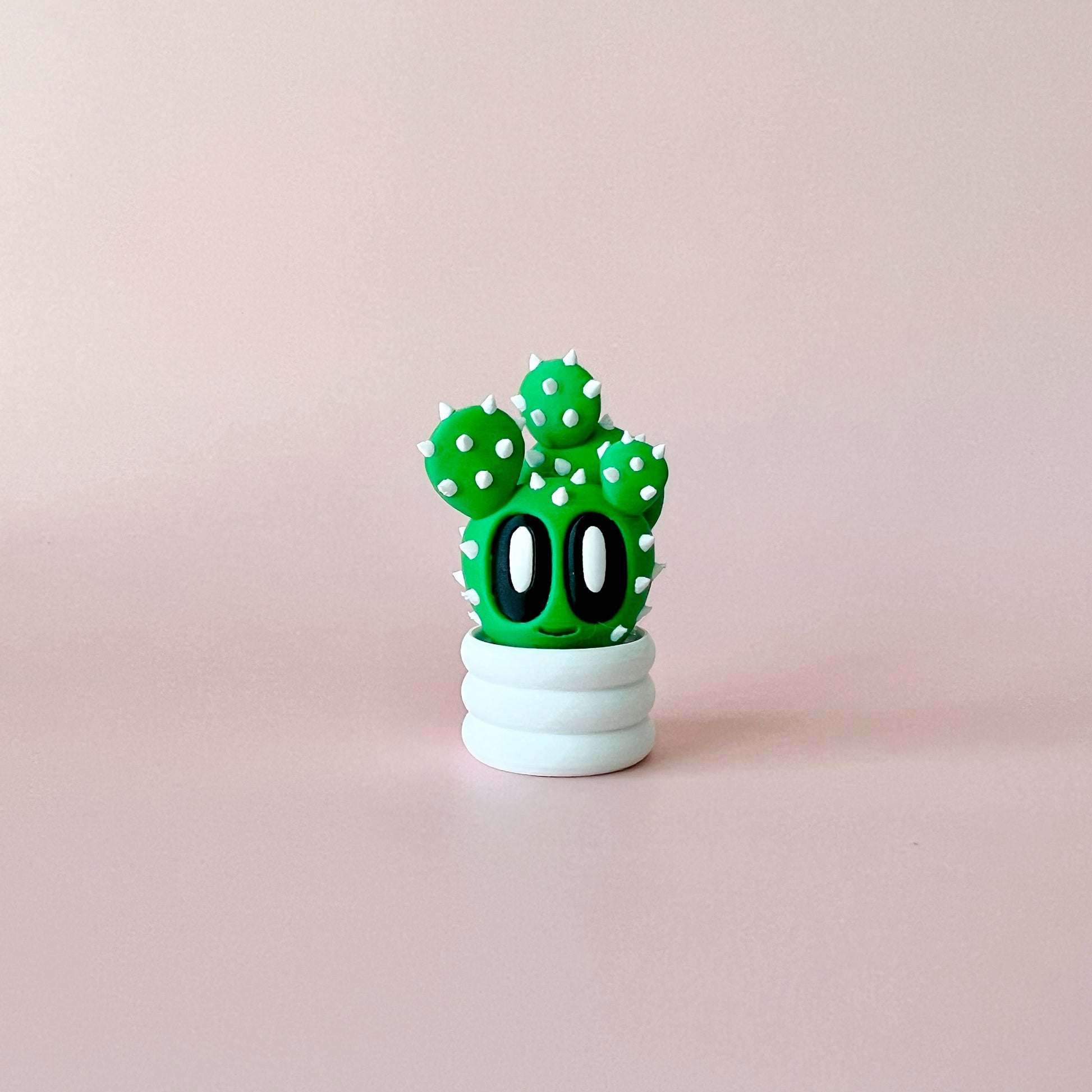 A whimsical green cactus with big eyes and white spikes, sitting in a round white pot against a soft pink background.