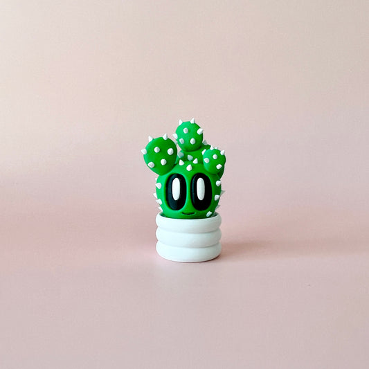 A whimsical green cactus with big eyes and white spikes, sitting in a round white pot against a soft pink background.