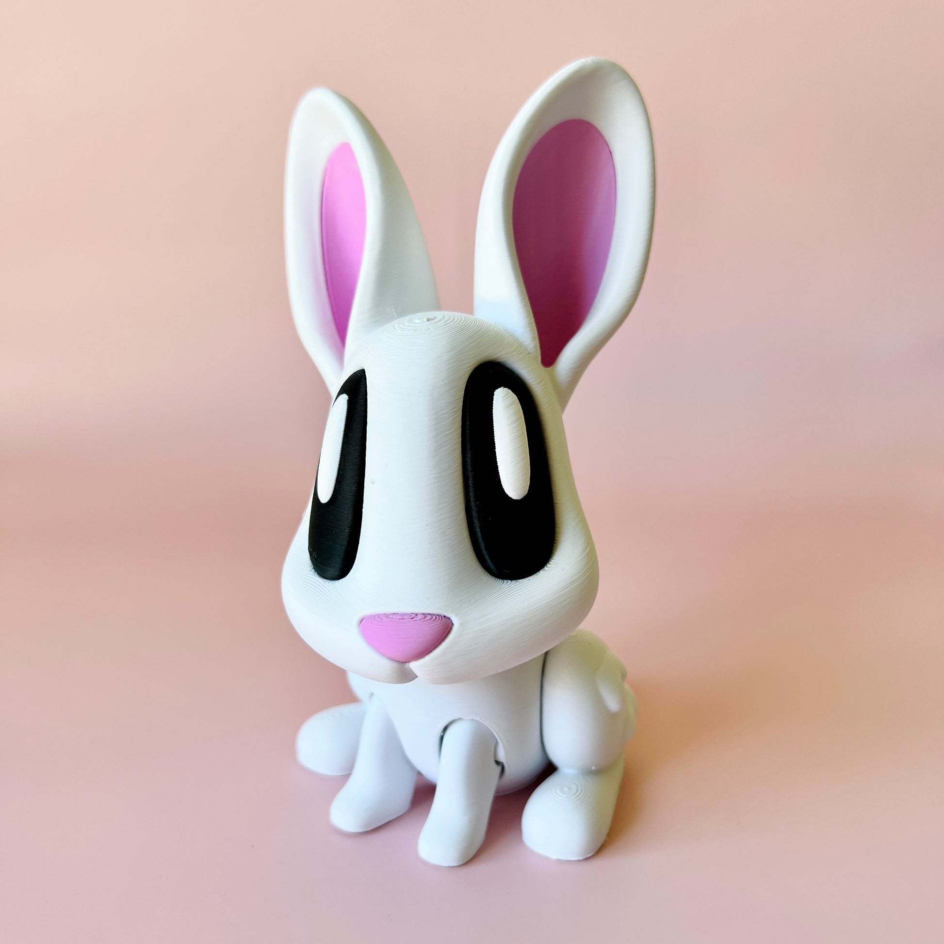 A cute, white bunny figure with large black eyes and pink inner ears, set against a soft pink background.