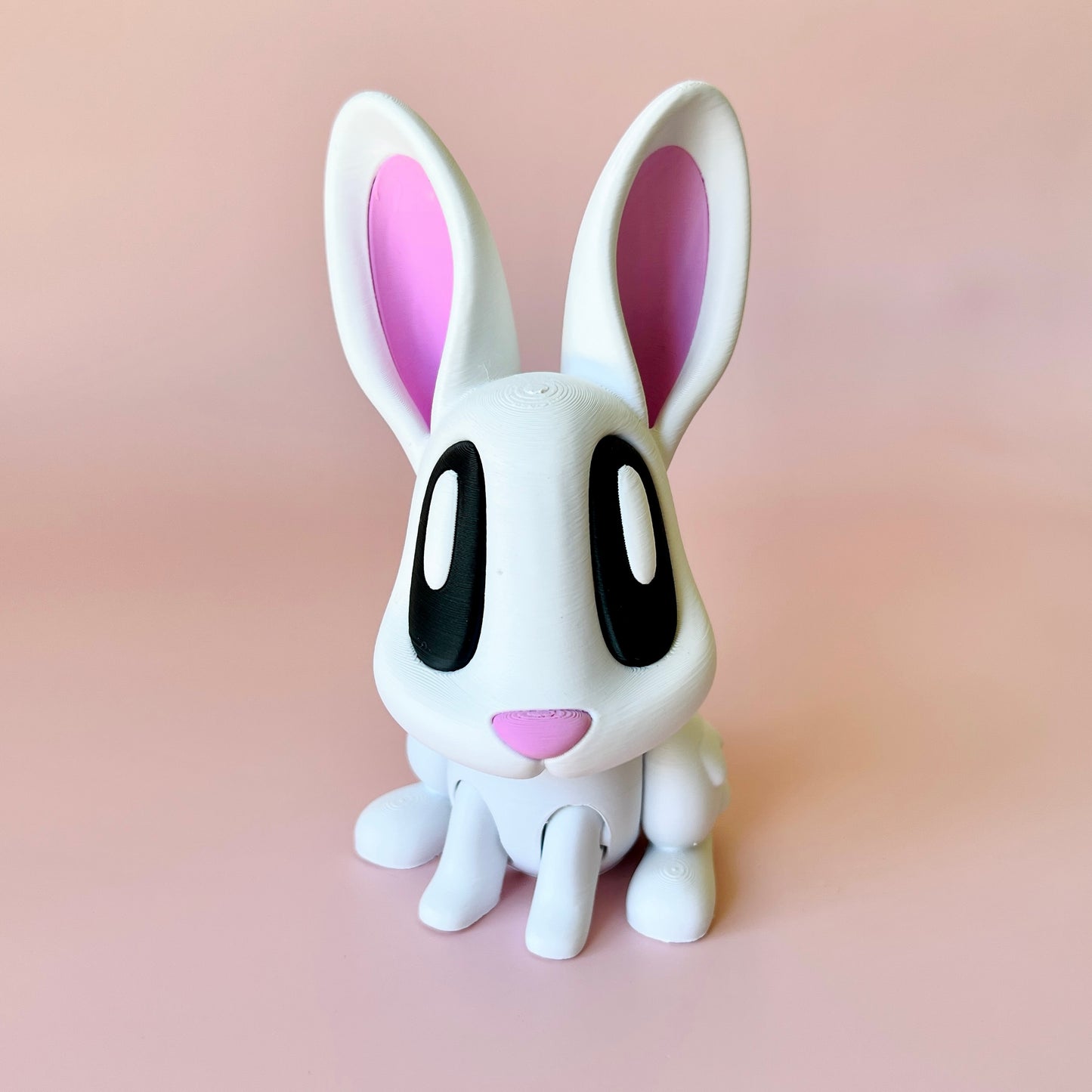 A whimsical white rabbit figure with pink accents and large black eyes, set against a soft pink background.