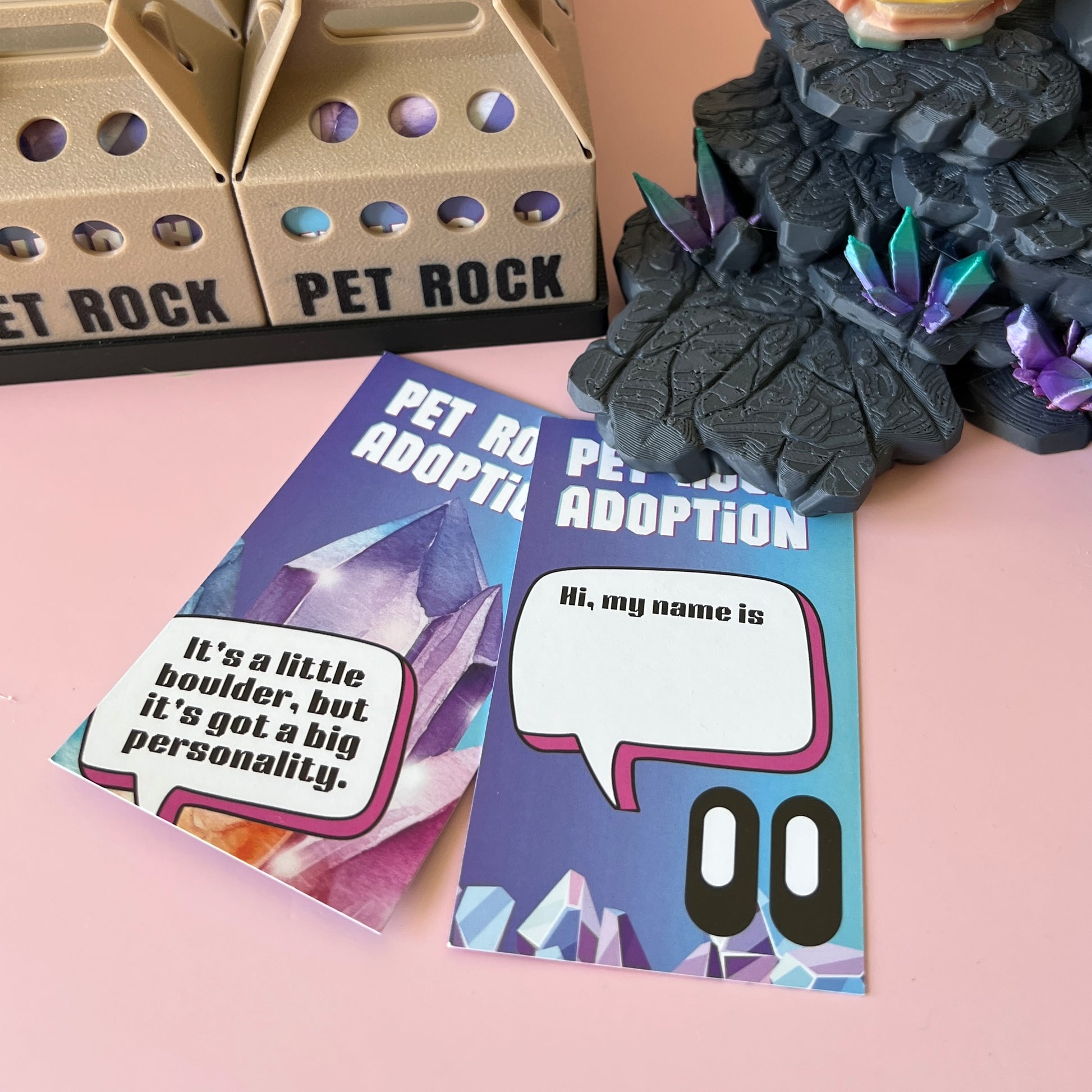 Two colorful adoption cards with playful text, displayed beside a decorative rock-themed display on a pink surface.