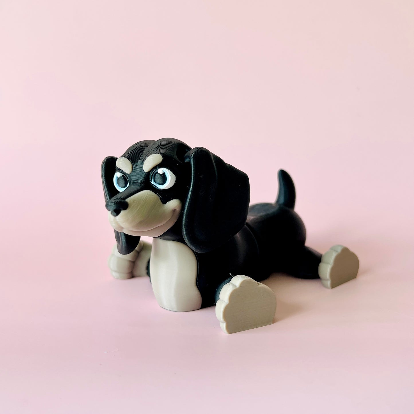 A playful black and beige dog figure with big blue eyes, lounging against a soft pink background.