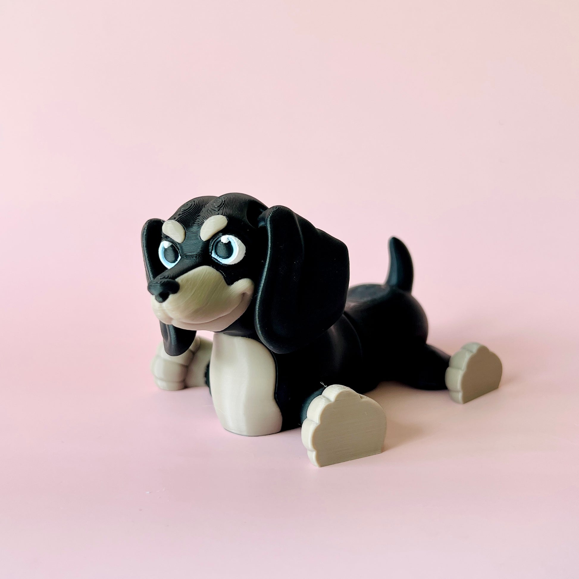 A playful black and beige dog figure with big blue eyes, lounging against a soft pink background.