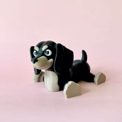 A playful black and beige dog figure with big blue eyes, lounging against a soft pink background.
