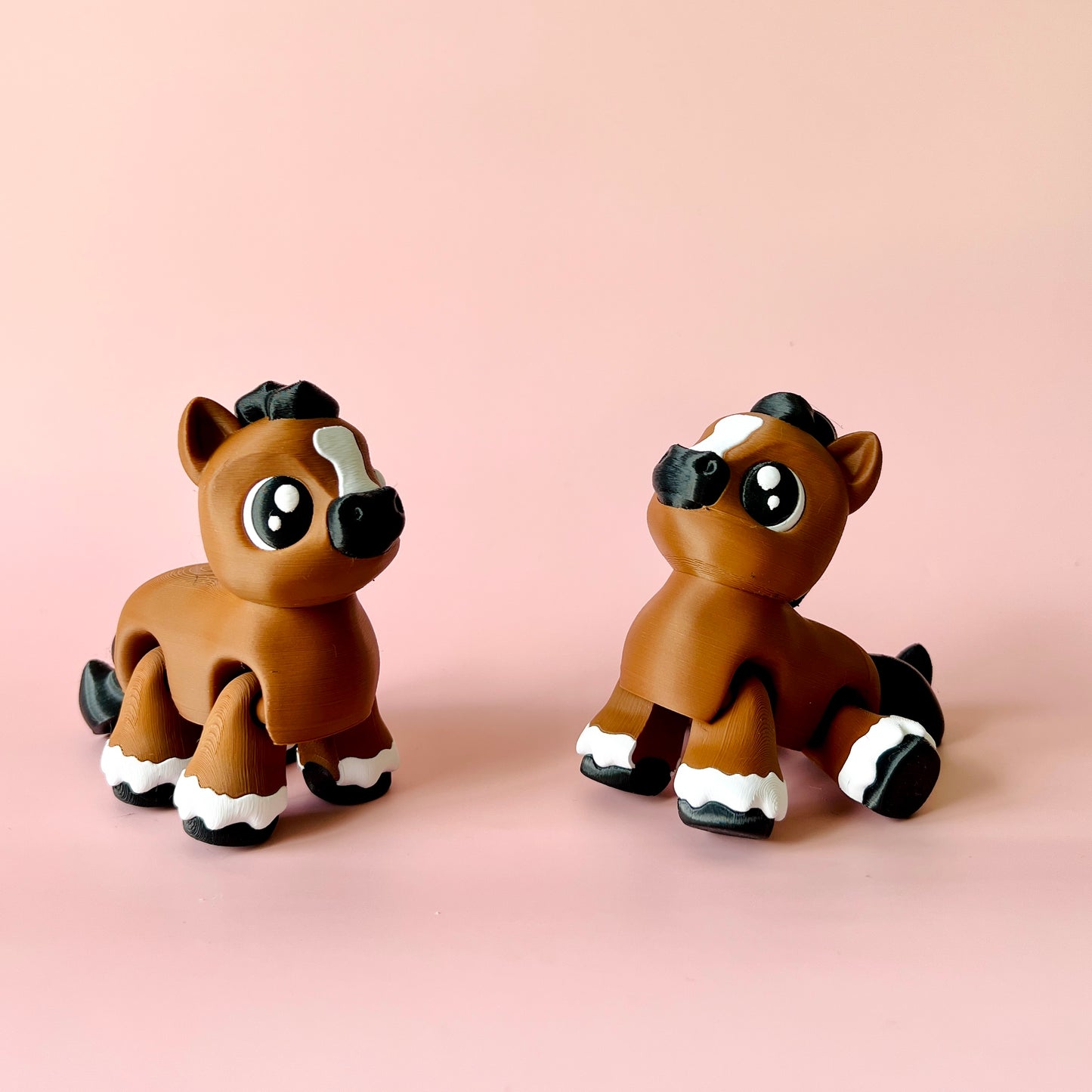 Two cute, stylized brown foals with large eyes and white accents, set against a soft pink background.