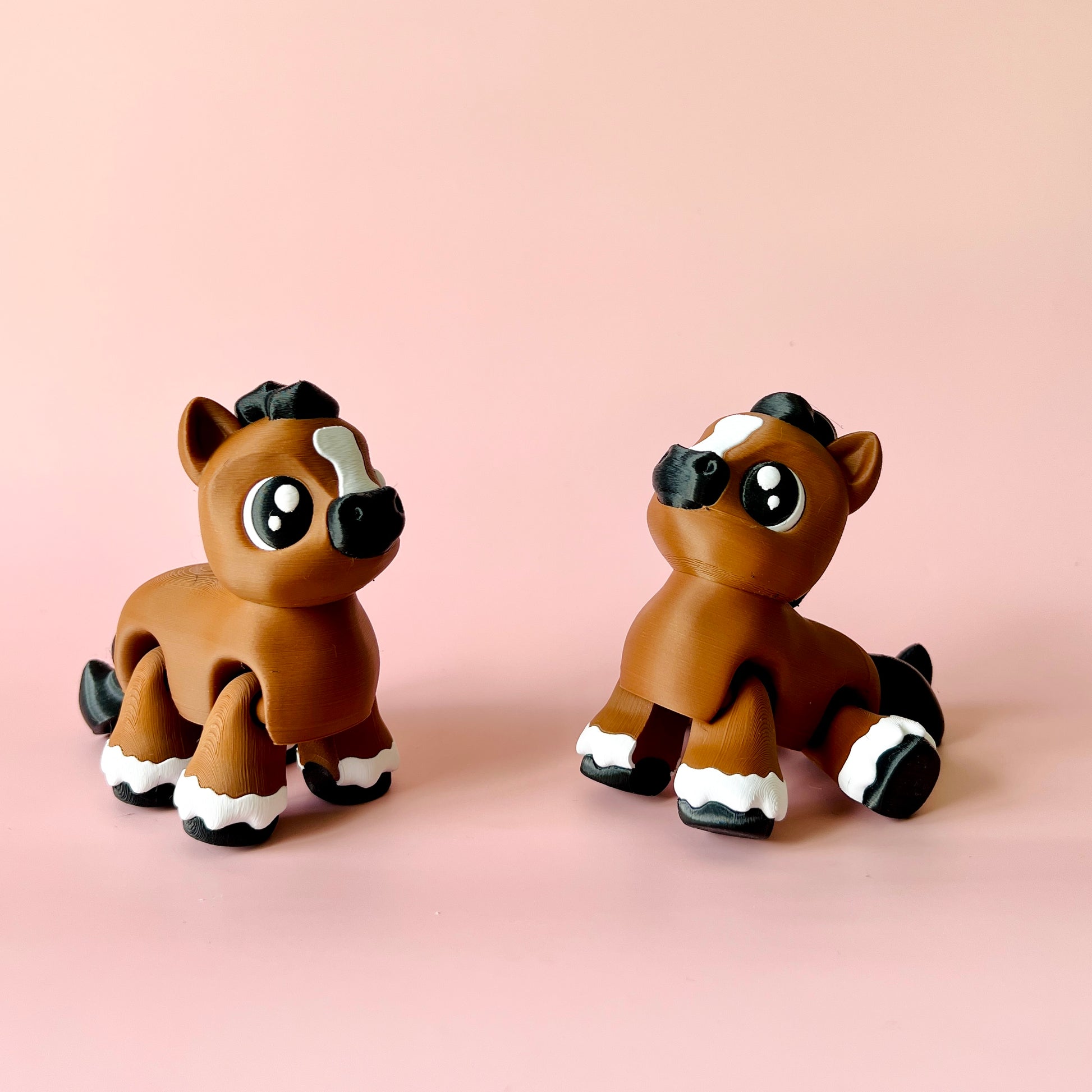Two cute, stylized brown foals with large eyes and white accents, set against a soft pink background.