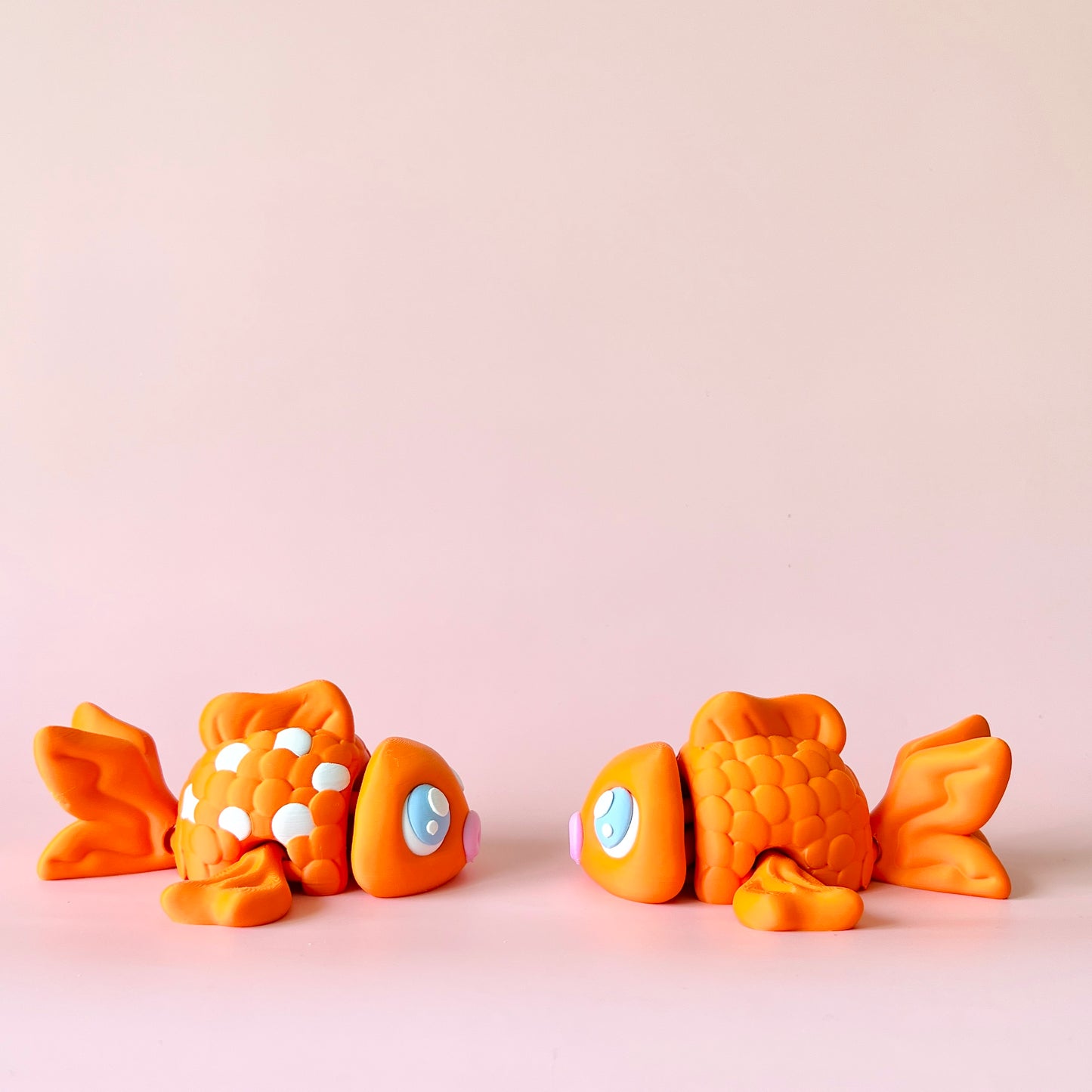 Two playful orange fish with bright blue eyes and varied textures on a soft pink background.