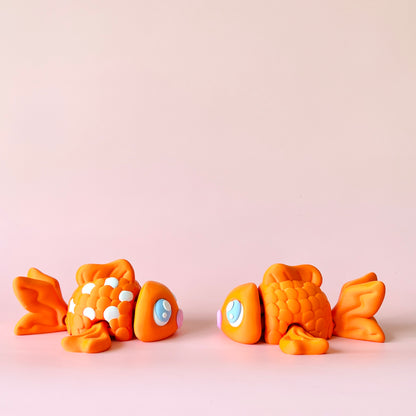 Two playful orange fish with bright blue eyes and varied textures on a soft pink background.