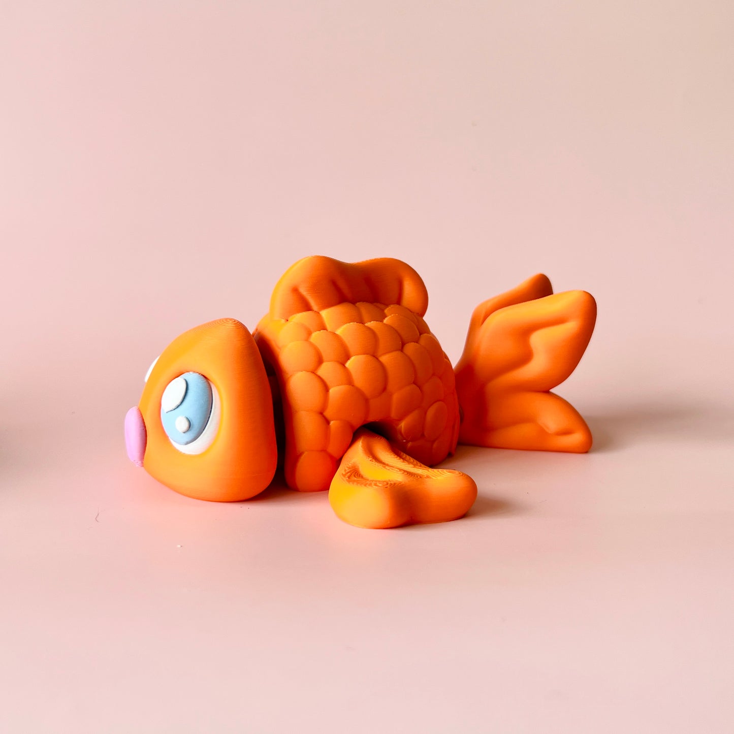 Vibrant orange fish with large blue eyes and a pink nose, resting on a soft pastel background.
